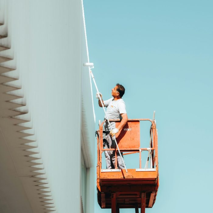 A man painting a building exterior from a cherry picker against a clear blue sky.